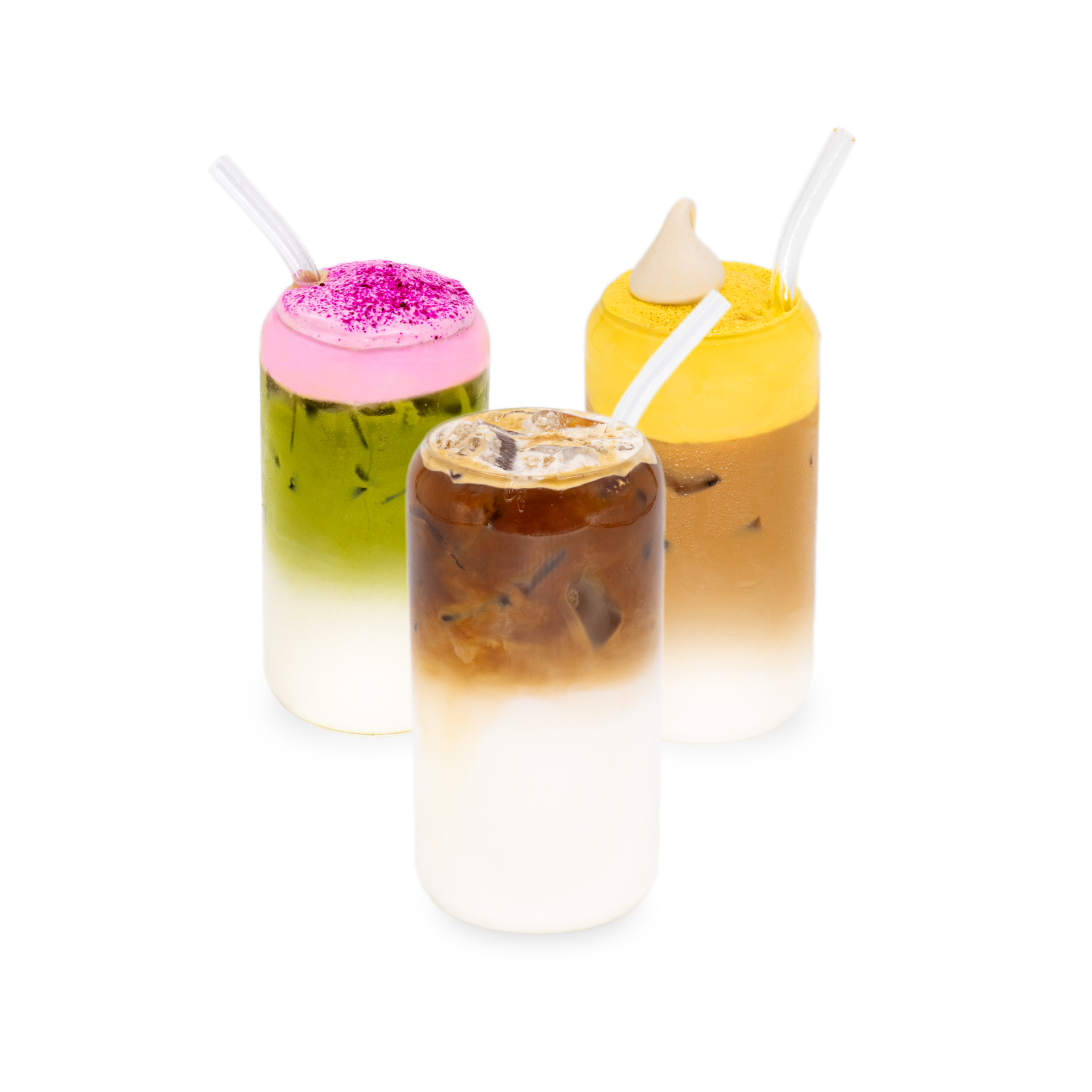 Three iced layered drinks in clear glasses with straws: a green and white drink topped with pink foam, a brown and white drink with ice, and a yellow and white drink with a dollop of tan cream.