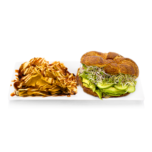 Croissant sandwich with avocado, cucumber, sprouts and chips drizzled with sauce on a white plate