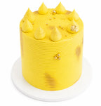 Tall lemon cake with bright yellow frosting, decorated with dollops, gold accents, and textured sides, presented on a white cake board.