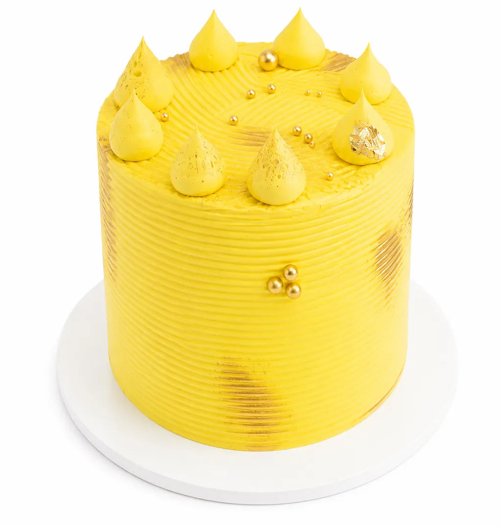 Tall lemon cake with bright yellow frosting, decorated with dollops, gold accents, and textured sides, presented on a white cake board.