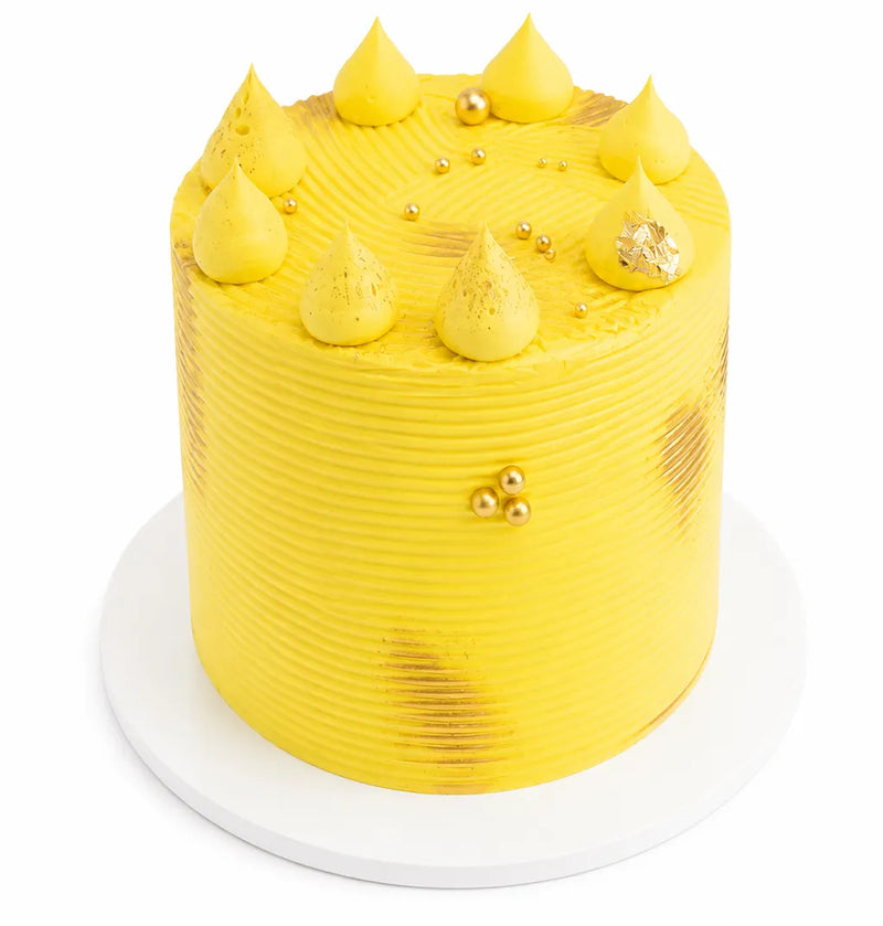 Tall lemon cake with bright yellow frosting, decorated with dollops, gold accents, and textured sides, presented on a white cake board.
