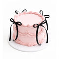 Pink Celebration Cake with smooth pale pink frosting, decorated with piped swirls and black ribbon bows around the sides for an elegant, custom birthday cake look.