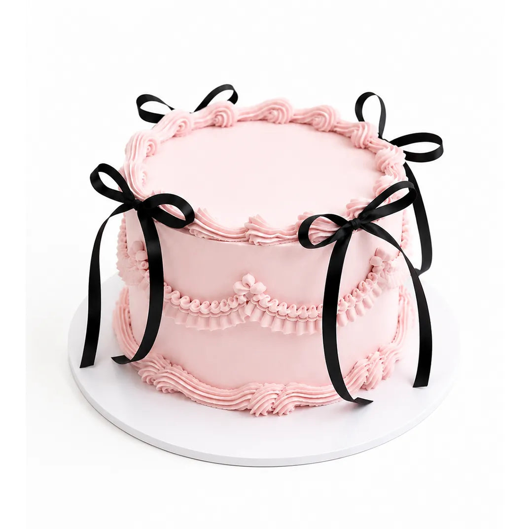 Pink Celebration Cake with smooth pale pink frosting, decorated with piped swirls and black ribbon bows around the sides for an elegant, custom birthday cake look.