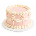 Velvet Whisper Cake featuring smooth light pink frosting with elegant white piped swags, rosettes, and scalloped borders on a white cake board.