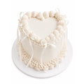 White Dream Heart Cake elegant white heart-shaped cake decorated with intricate piped frosting borders and delicate white ribbon accents on a round base.