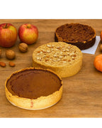 Three gourmet pies with almond, apple, and pumpkin, surrounded by apples, nuts, and pumpkins on wood table.