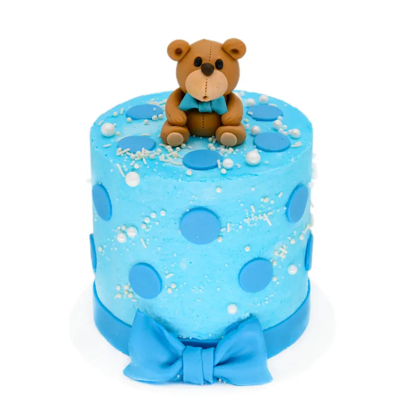 Baby Boy Cake featuring a blue frosted design with large blue polka dots, white sprinkles, a fondant bow at the base, and a sitting teddy bear topper for a charming celebration.