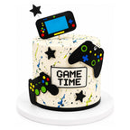 Round white cake with colorful paint splatter design featuring fondant video game controllers and a handheld console with "Game Time Cake" text in pixelated font on the front.