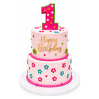 Two-tier pink birthday cake decorated with small colorful flowers and green buttons, topped with a large pink number one and gold "Happy Birthday" lettering on a white base.
