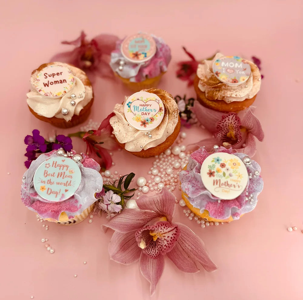 Six decorated cupcakes with Mothers Day messages sit on a pink surface, surrounded by pink flowers and pearl-like beads. Each cupcake has colorful frosting and a unique decorative topper.