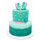 Two-tier aqua-themed cake featuring wavy ocean-like layers on the bottom tier, a "6" in gold on the upper tier, decorated with shells, stars, and a pair of teal and pink flip-flop sandals on top