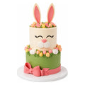 Two-tier cake decorated as a bunny with pink ears, closed eyes, and a pink nose, surrounded by pastel-colored flowers and a pink bow on the green lower tier.