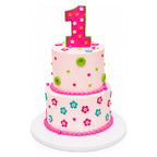 Two-tiered pink birthday cake decorated with colorful small flowers, green buttons, and tiny hearts, topped with a large pink number one outlined in green.