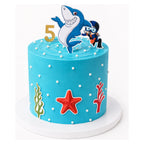 Ocean Adventure Cake with blue icing decorated with white dots, seaweed, a red starfish, a gold number 5, and toppers of a smiling shark and a scuba diver.