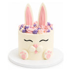 Cute Bunny Cake featuring a white fondant exterior with pink and purple floral decorations on top, bunny ears, closed eyes with lashes, and pink paw prints on the front.