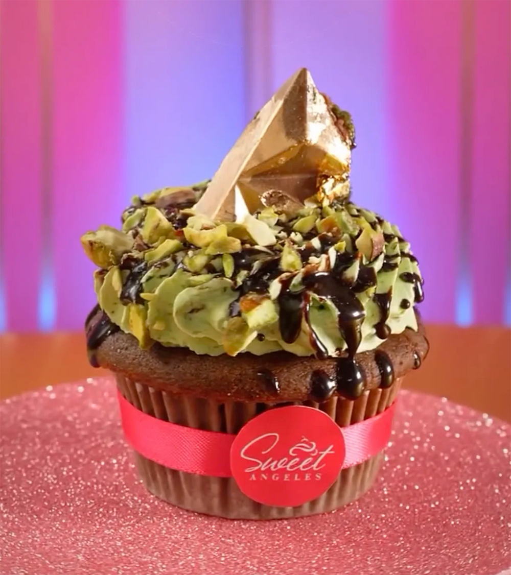 A chocolate cupcake topped with green frosting, chopped nuts, chocolate drizzle, and a gold-tipped geometric chocolate piece sits on a pink glittery surface with a “Sweet Angeles” label, against a pink and purple background.